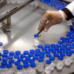 Gloved technician handling vials on a pharmaceutical vaccine production line, representing API and drug supply chains exposed to tariff risk