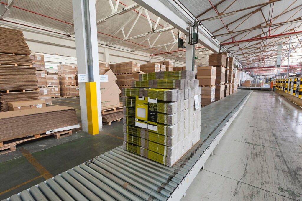 Palletised corrugated shipping boxes on a roller conveyor in a consumer goods packaging warehouse, illustrating AI supply chain risk management for packaging suppliers