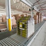 Palletised corrugated shipping boxes on a roller conveyor in a consumer goods packaging warehouse, illustrating AI supply chain risk management for packaging suppliers